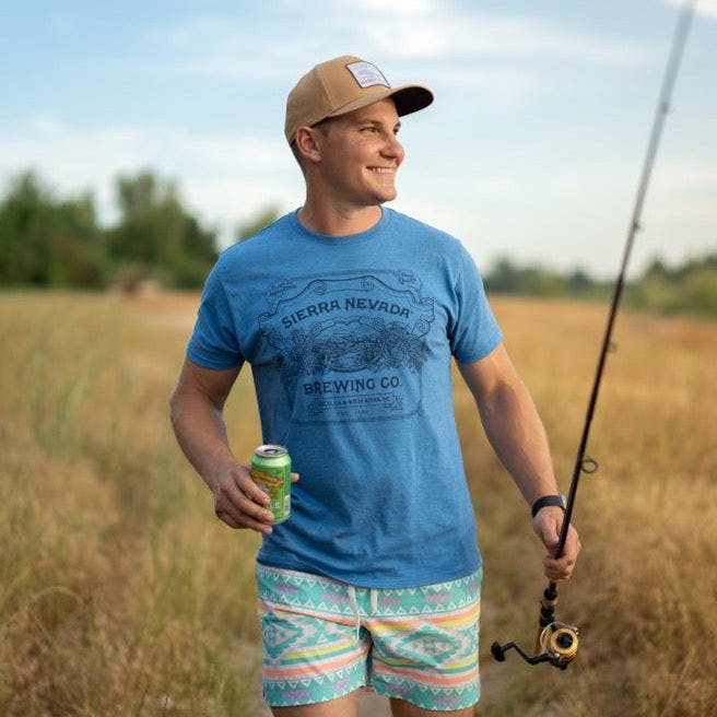 A man wears the Sierra Nevada Brewing Co. Handcrafted T-Shirt while walking along a riverbank carrying a fishing pole and a Pale Ale.