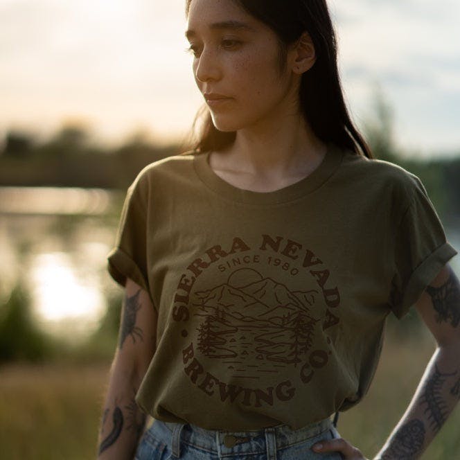 A woman wears the Sierra Nevada Brewing Co. Mountain Circle T-Shirt while standing near a peaceful river.