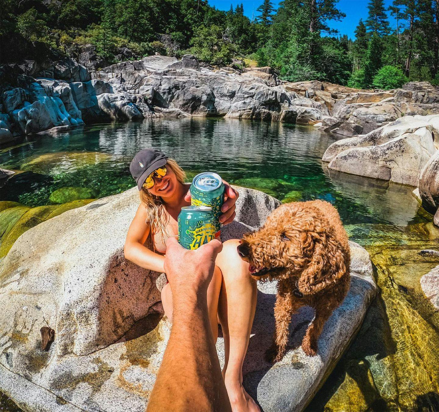 Woman with a dog sitting in the sunshine on a rock next to a lake while toasting someone with a can of Hazy Little Things IPA beer